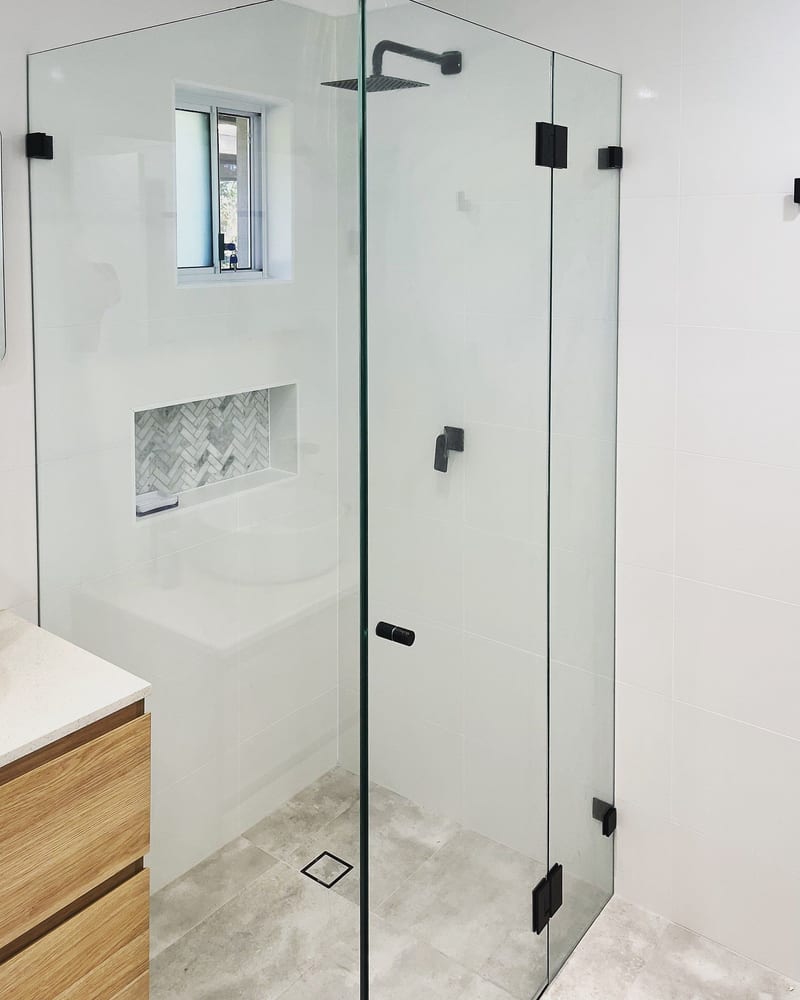 Clear frameless corner shower screen with hinge panel, door and return panel in black hardware, white wall tiles and a timber vanity
