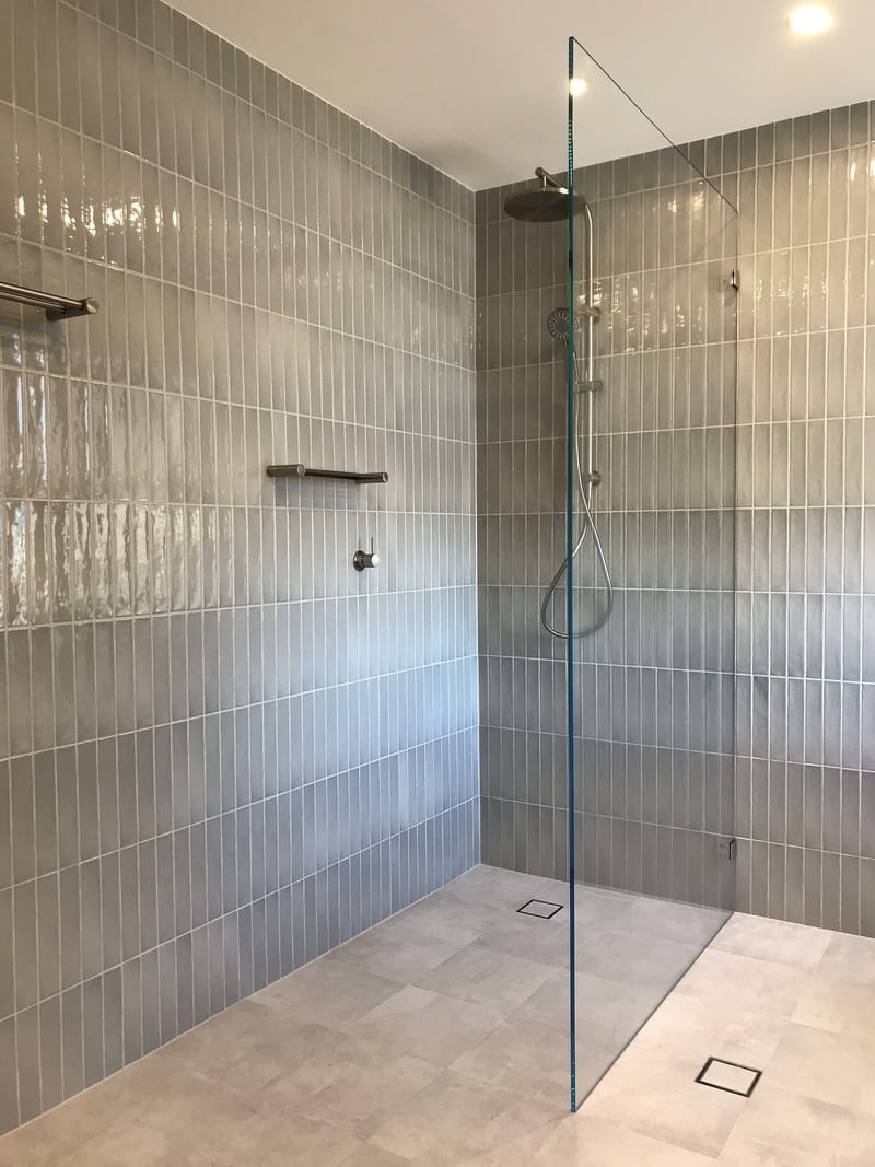 Walk-in frameless fixed panel against grey vertical tiles with a pale tiled floor